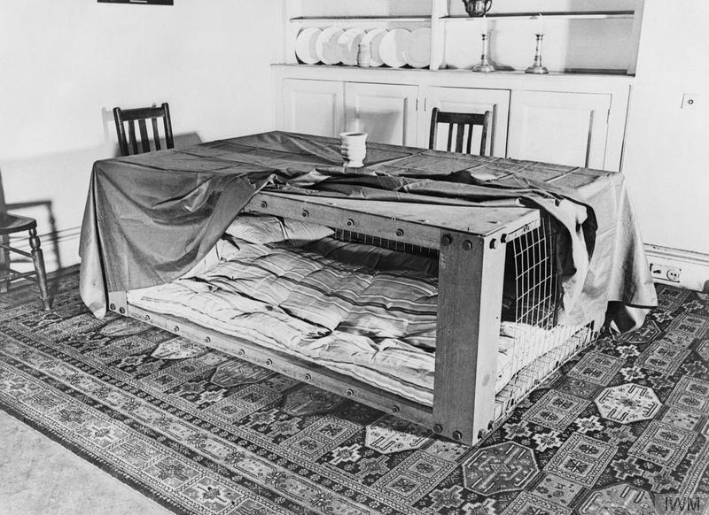 A photograph of a Morrison shelter in a room setting, showing how such a shelter could be used as a table during the day and as a bed at night. © IWM (D 2053)